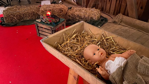 In einer mit Stroh ausgelegten Krippe liegt eine Babypuppe. Im Hintergrund liegen rechteckige Strohballen auf einem Holzboden. © NDR Foto: Wieland Gabcke