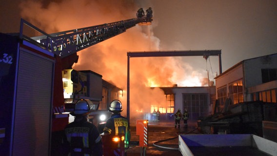 Mehrere Feuerwehrleute stehen vor einer Lagerhalle, die lichterloh brennt. © FW Gifhorn Foto: FW Gifhorn