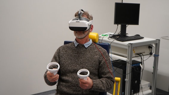 Ein Mann hat eine VR-Brille auf dem Kopf und entsprechende Controller in den Händen. © UMG Universitätsmedizin Göttingen Foto: Lukas Diedrich