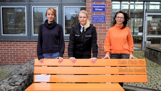 Polizeipräsidentin Tanja Wulff-Bruhn (Mitte) mit den Gleichstellungsbeauftragten der Polizeidirektion Göttingen Stefanie Wattenberg (links) und Dörte Köhler (rechts) vor einer orangenen Bank, die auf Gewalt gegen Frauen hinweist. © Polizeiinspektion Göttingen 