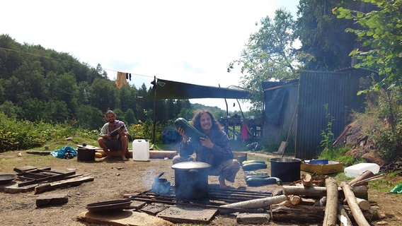 Ein Mann sitzt vor einem Kochtopf auf einem Grillplatz und hält eine große Zucchini in der Hand. © NDR Foto: Marco Schulze