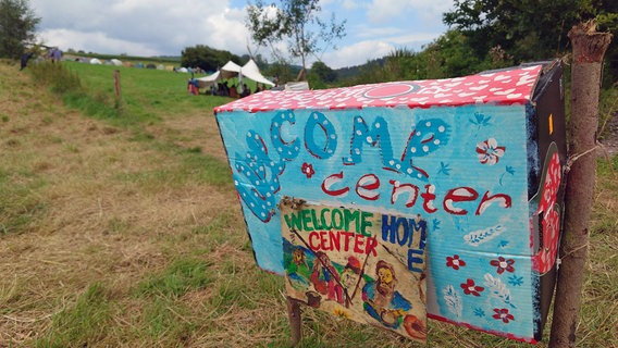 Auf einem bunt bemalten Karton vor einer Wiese mit Zelten steht die Aufschrift "Welcome Center" © NDR Foto: Marco Schulze