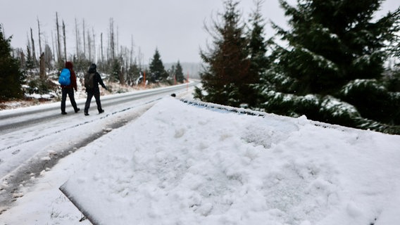 Zwei Wanderer laufen über einen schneebedeckten Waldweg. Im Vordergrund ist eine schneebedeckte Hinweistafel zu sehen. © dpa Foto: Matthias Bein