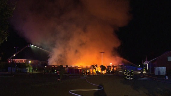 Auf dem Foto ist es Nacht, im Hintergrund ist ein großer Feuerschein zu sehen. Vorne stehen mehrere Einsatzkräfte der Feuerwehr und löschen, ein Schlauch liegt auf dem Boden. © Hannover Reporter 