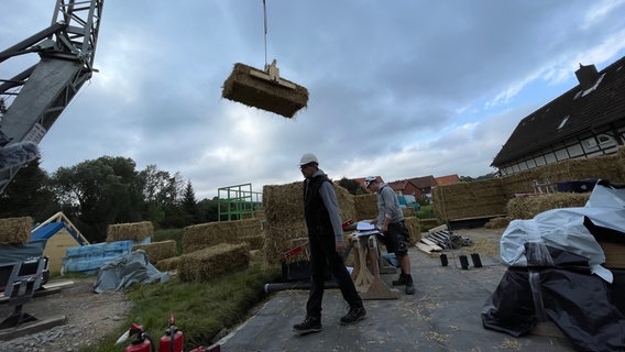 Ein Zimmerer auf einer Strohhausbaustelle, über ihm schwebt ein Strohballen an einem Kran. © NDR Foto: Jan Fragel