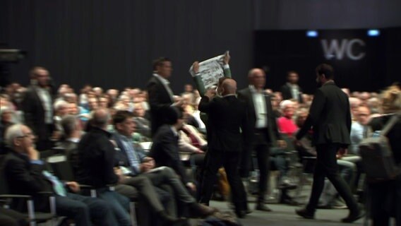 Sicherheitspersonal eskortiert einen Mann nach einem Tortenwurf aus dem Saal, in welchem die VW Hauptversammlung statt findet. © NDR 