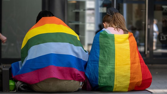 Teilnehmende des CSD sitzen in Fahnen gehüllt auf einer Bank. © picture alliance/Flashpic/Jens Krick Foto: Jens Krick