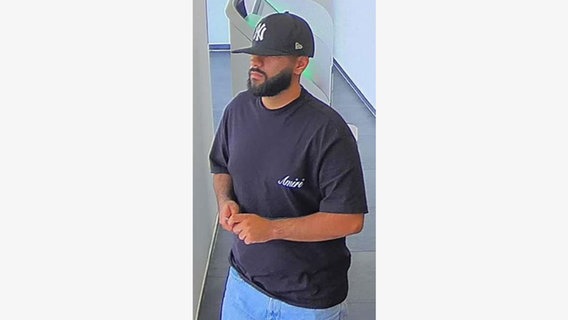 Ein Mann mit dunklem Bart, schwarzer Cap mit weißer Aufschrift, schwarzem T-Shirt und heller Jeans. © Pressestelle Polizei Bremen 