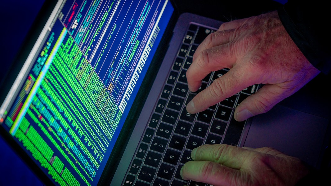 Symbolbild zu Computerhackern. Hände an einem Laptop. Auf dem Bildschirm ist grüne Schrift zu sehen.