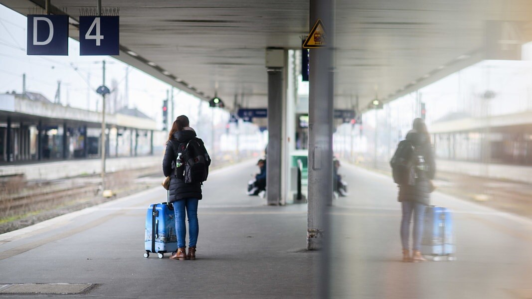 Eine Frau wartet mit einem Koffer am Gleis 4 im Hauptbahnhof Hannover und spiegelt sich in einer Glasscheibe.