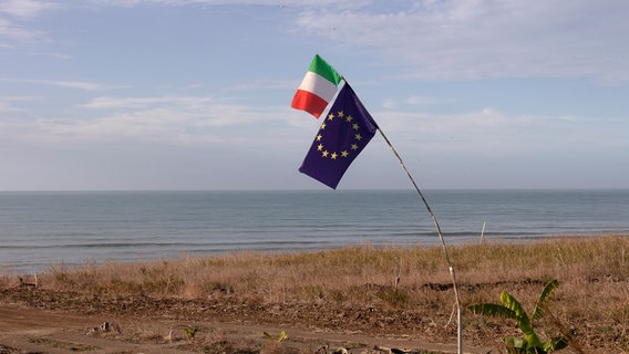 Ein Mast mit einer italienischen und einer europäischen Flagge steht gebeugt auf einer vertrockneten Wiese vor dem Meer. © Hochschule Hannover Foto: Nora Börding und Anne Speltz