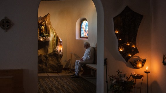 Ein Geistlicher sitzt in einer kleinen, dunklen Kapelle auf einer Bank. © Hochschule Hannover Foto: Marcel Zeumer