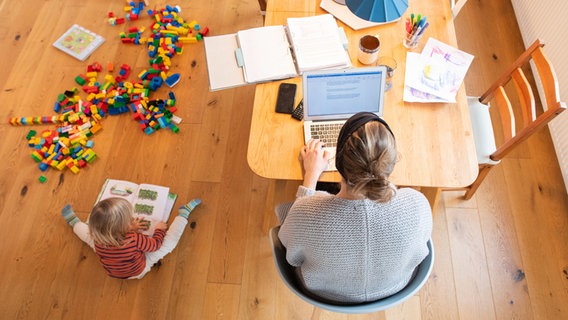 Ein zweijähriges Kind spielt im Wohnzimmer, während seine Mutter im Homeoffice an einem Laptop arbeitet. © dpa-Bildfunk Foto: Julian Stratenschulte