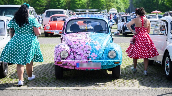 Zwei Frauen in Kleidern laufen neben einem Volkswagen Käfer im Blümchenoptik beim MaiKäfertreffen auf dem Messegelände. © picture alliance/dpa | Julian Stratenschulte Foto: Julian Stratenschulte