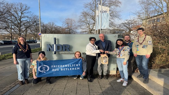 Vor dem NDR-Schild stehen Pfadfinder und halten ein Transparent mit der Aufschrift "Friedenslicht aus Betlehem", ein Mädchen übergibt Funkhausdirektorin Andrea Lütke ein Friedenslicht. © NDR Foto: Marco Heuer