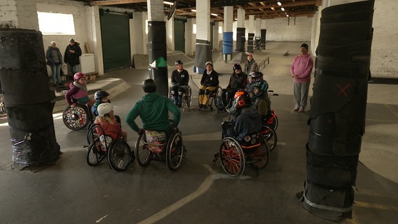 Eine Gruppe Menschen in Rollstühlen sitzen beieinander in einer Halle, in der sie skaten. © NDR 