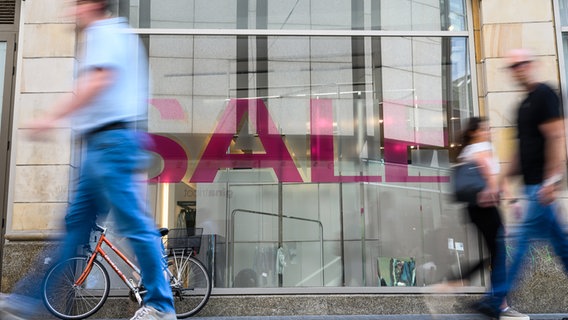 Menschen gehen an einem Schaufenster vorbei in dem ein Schild mit der Aufschrift "Sale" hängt. © dpa-Bildfunk Foto: Christophe Gateau/dpa