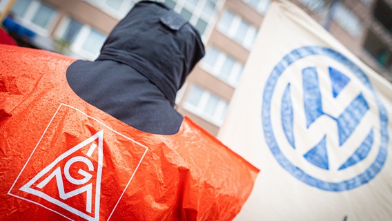 Ein Mann steht mit einer roten Weste auf der das IG-Metall-Lgo zu sehen ist vor einem VW-Banner. © picture alliance Foto:  | Moritz Frankenberg