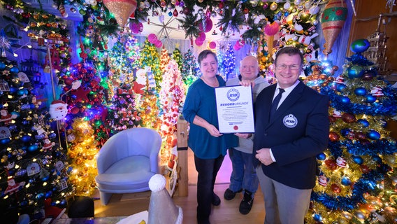 Susanne und Thomas Jeromin stehen mit Olaf Kuchenbecker (r) vom Rekord-Institut für Deutschland inmitten von Weihnachtsbäumen in ihrem Haus im Landkreis Schaumburg. © Julian Stratenschulte/dpa Foto: Julian Stratenschulte