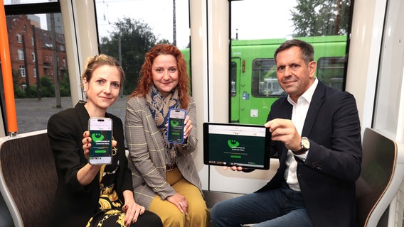 Wirtschaftsminister Olaf Lies, die Üstra-Vorsitzende und eine Vorständin der Üstra halten Handys und Tablets in die Kamera während sie in einer Stadtbahn sitzen. © ÜSTRA/Florian Arp 
