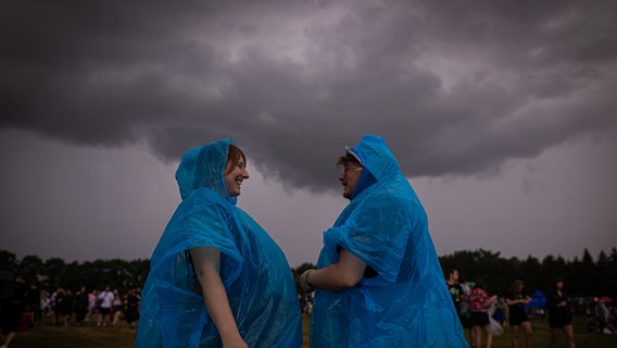 Zwei Festivalbesucher tragen Regenponchos, während dunkle Regenwolken über das Gelände des Hurricane Festivals in Scheeßel hinwegziehen. © dpa Foto: Moritz Frankenberg