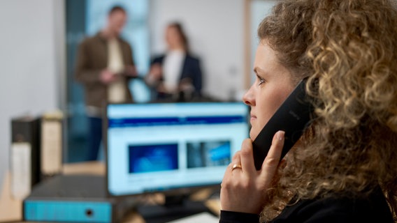 Eine junge Frau telefoniert in einem Büro. Im Hintergrund sind unscharf zwei weitere Mitarbeitende zu sehen. © NDR 