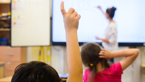 Ein Grundschüler meldet sich per Handzeichen, während eine Lehrerin auf eine digitale Schultafel in einem Klassenraum schreibt. © picture alliance / dpa | Julian Stratenschulte Foto: Julian Stratenschulte