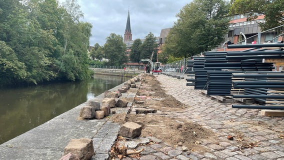 Auf dem gepflasterten Weg, der am Ufer eines Gewässers entlang führt, liegen viele Metallstangen. An einigen Stellen nah am Wasser hat das Pflaster Löcher: Die Steine, die dafür aus dem Kopfsteinpflaster entfernt wurden, liegen auf dem Weg. © Stadt Lüneburg 