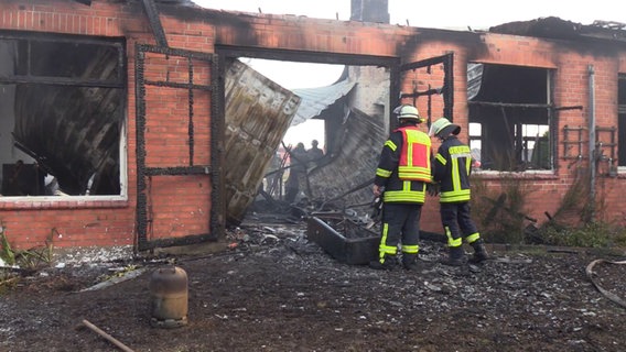 Einsatzkräfte der Feuerwehr stehen vor einer abgebrannten Halle. © NonstopNews 