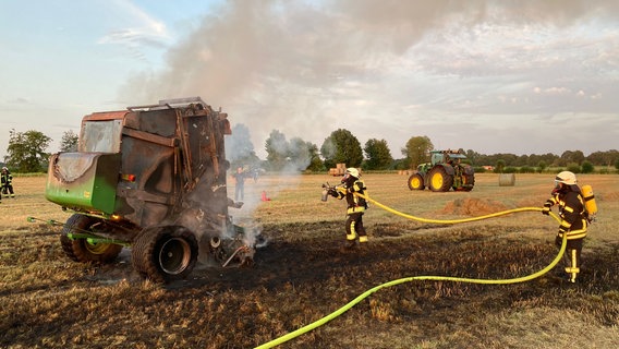 Zwei Feuerwehrleute in Einsatzkleidung mit Atemschutz löschen mit einem Schlauch eine brennende Ballenpresse auf einem Feld. © Polizei Stade 
