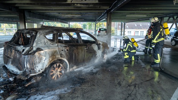 Auf dem Foto ist ein ausgebranntes Auto zu sehen, das auf einem Parkdeck steht. Rechts daneben befinden sich drei Feuerwehrleute, die mit Löscharbeiten beschäftigt sind. Aus einem Schlauch spritzen sie Wasser auf das Auto. © Joto - Tobias Johanning Foto: Tobias Johanning