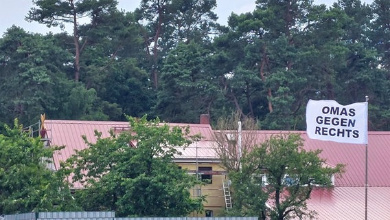 Bei einer Demo gegen Rechtsextremismus in Eschede im Landkreis Celle steht "Omas gegen rechts" auf einer Fahne, im Hintergrund ist ein Gelände zu sehen, auf dem kürzlich Neonazis zusammengekommen sind. © NDR Foto: Julia Scheper