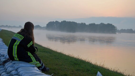 Eine Frau sitzt auf Sandsäcken auf einem Deich und schaut auf das Wasser. Baumkronen ragen heraus. Nebel zieht über das Wasser. © NDR Foto: Sven Lehmann