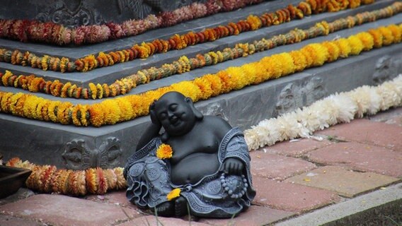 Die Figur eines Buddhas ist mit gelben Blumen verziert. Im Hintergrund liegen weitere Blumenketten. © NDR Foto: Vivian Münzel