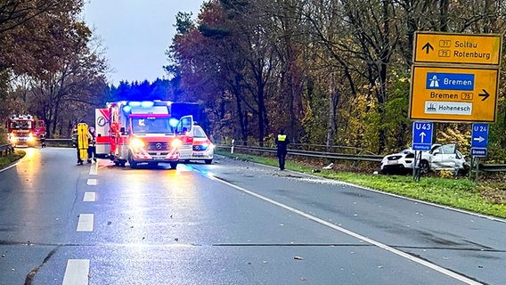 Ein stark beschädigtes Unfallfahrzeug steht im Grünstreifen der Bundesstraße, während auf der Fahrbahn mehrere Einsatzwagen der Feuerwehr und des Rettungsdienstes stehen. © Polizeiinspektion Rotenburg 