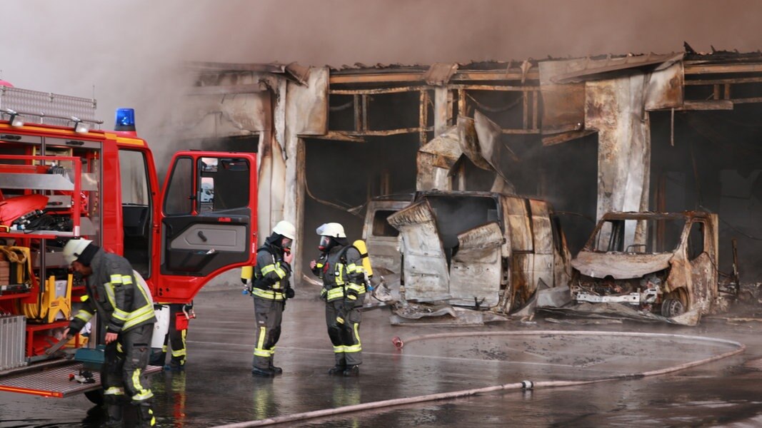 Ausgebrannte Autos und Einsatzkräfte der Feuerwehr stehen vor einem brennenden Autohaus.