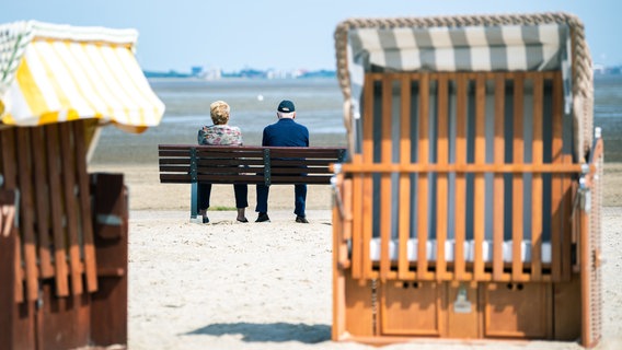 Zwei Personen sitzen auf einer Bank am Strand, im Vordergrund Strandkörbe. © dpa Bildfunk Foto: Mohssen Assanimoghaddam