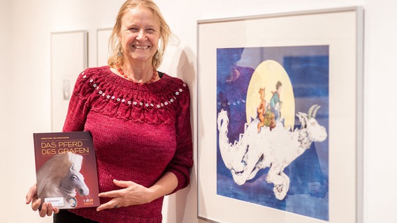 Cornelia Funke, deutsche Kinder- und Jugendbuchautorin, steht mit dem Buch "Das Pferd des Grafen" in der Kinderbibliothek am Kulturzentrum PFL neben einem Bild. © dpa Foto: Hauke-Christian Dittrich