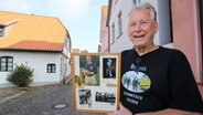 Ein Mann mit grauen Haaren steht lächelnd mit einem Bilderrahmen in der Hand vor einer Hauswand. © dpa-Bildfunk Foto: Focke Strangmann