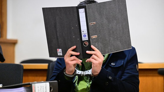 Ein Angeklagter bedeckt in einem Gerichtssaal sein Gesicht mit einem Aktenordner. © dpa Foto: Sina Schuldt