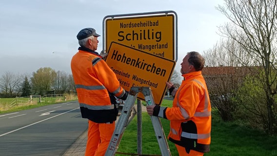 Zwei Männer Tauschen zwei Ortsschilder mit den Namen "Schillig" und "Hohenkirchen" aus. © NDR Foto: Jutta Przygoda