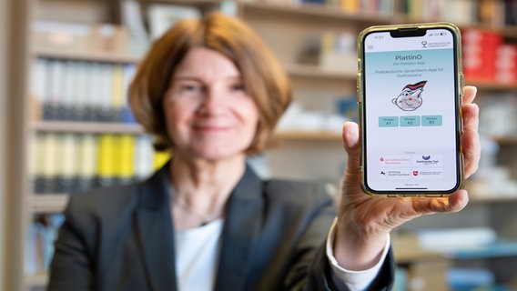 Eine Frau hält ein Smartphone mit ausgestrecktem Arm in eine Kamera, darauf ist die App "PlattinO" zu erkennen. © Ostfriesische Landschaft 