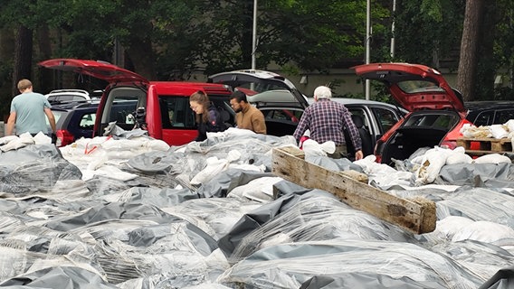 Fahrzeuge mit geöffneten Kofferraumen und einige Personen, im Vordergrund ein riesiger Haufen mit Plastik abgedeckter Sandsäcke. © NDR Foto: Sebastian Duden