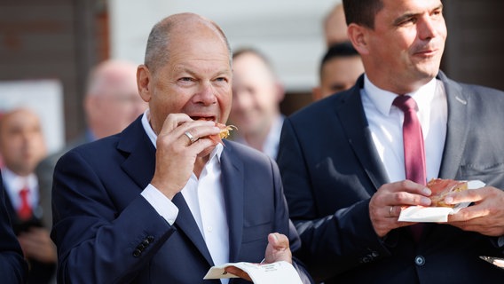 Bundes·kanzler Olaf Scholz isst ein Schinken-Brot auf dem Stoppel·markt in Vechta. Neben Olaf Scholz steht Kristian Kater. Kristian Kater ist der Bürger·meister von Vechta. © dpa-Bildfunk Foto: Friso Gentsch