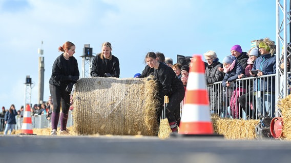 Der Strohballen wird von einem Frauenteam den Deich hinaufgerollt. Zahlreiche Zuschauer verfolgen die 13. Ostfriesische Strohballen-Rollmeisterschaft, bei der Männer-, Frauen- und Mixed-Teams um den Titel "Strohballen-Rollmeister 2024" wetteifern. © Lars Penning/dpa Foto: Lars Penning