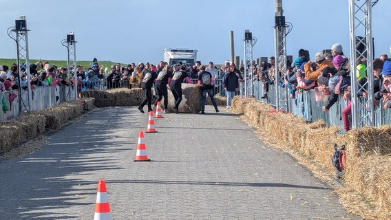 Der Strohballen wird von einem Frauenteam den Deich hinaufgerollt. Zahlreiche Zuschauer verfolgen die 13. Ostfriesische Strohballen-Rollmeisterschaft, bei der Männer-, Frauen- und Mixed-Teams um den Titel "Strohballen-Rollmeister 2024" wetteifern. © NDR Foto: Sebastian Duden