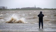 Ein Mann steht an einer Kaimauer und blickt auf das vom Sturm aufgepeitschte Wasser. © dpa-Bildfunk Foto: Sina Schuldt/dpa