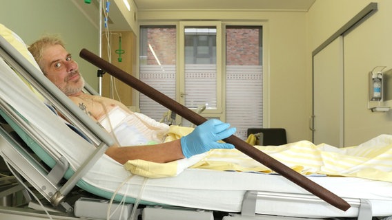 Patient Frank Vaorin liegt im Krankenhausbett und hält die Zaunstange in der Hand, die seinen Körper durchbohrte. © picture alliance/Gesundheit Nord · Klinikverbund Bremen Foto: Kerstin Hase
