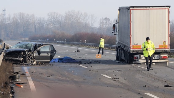 Auf einer Autobahn steht ein beschädigter Wagen mit öffenen Türen, daneben ein Lkw. Zwei Menschen in Warnkleidung laufen auf der Straße. © picture alliance/dpa Foto: Jörn Hüneke