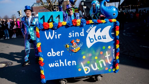 Akteure ziehen mit einem Schild "Wir sind zwar blau, aber wählen bunt" beim traditionellen Dammer Carnevalsumzug durch die Innenstadt. © Hauke-Christian Dittrich/dpa Foto: Hauke-Christian Dittrich/dpa